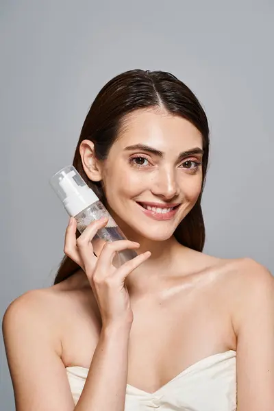A young Caucasian woman with brunette hair and clean skin, dressed in a white gown, gracefully holding a cleanser bottle. — Stock Photo
