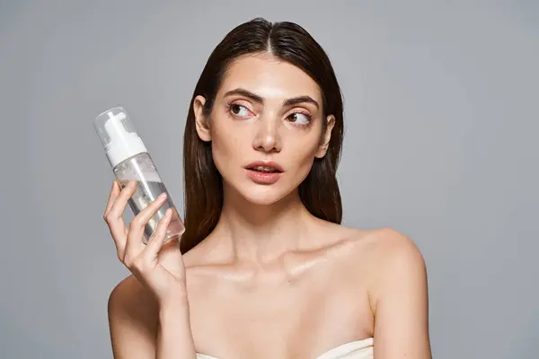 Una joven mujer caucásica con el pelo moreno sostiene una botella con limpiador facial de espuma delante de su cara, mostrando la piel limpia. - foto de stock