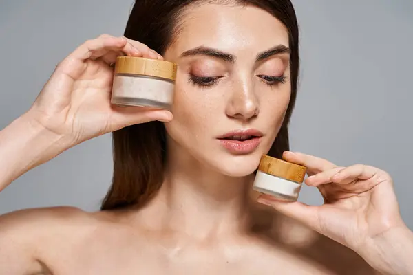 Jeune femme caucasienne aux cheveux bruns tenant deux pots de crème près de son visage pour nourrir et hydrater sa peau. — Photo de stock