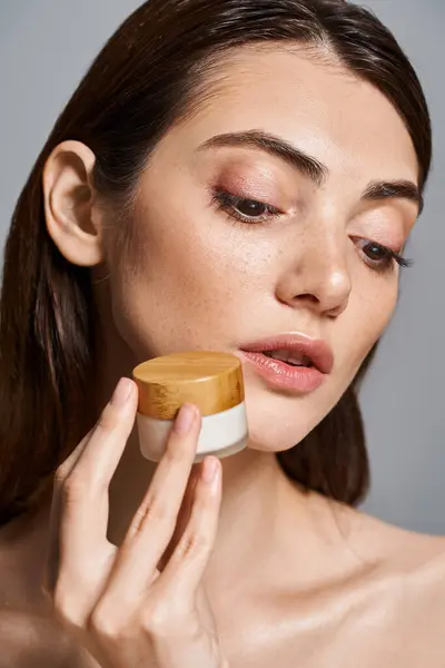 A young Caucasian woman with brunette hair holds a jar of cream to her face, gently applying the luxurious product. — Stock Photo