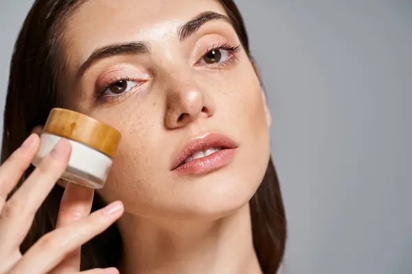Une jeune femme caucasienne aux cheveux bruns appliquant de la crème d'un bocal sur son visage, améliorant son éclat de peaux. — Photo de stock