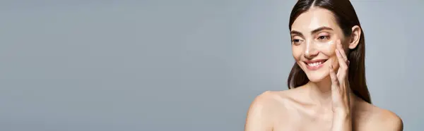 Una giovane donna caucasica con i capelli castani che sorride gioiosamente toccandosi delicatamente il viso, applicando uno striscione crema — Foto stock