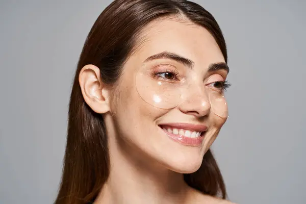 A young Caucasian woman with brunette hair smiles warmly, showcasing her clear and radiant skin with under eye patches — Stock Photo