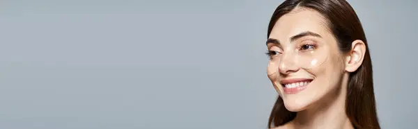 Une jeune femme caucasienne aux longs cheveux bruns sourit vivement à la caméra dans un décor de studio, sous la bannière des patchs oculaires — Photo de stock