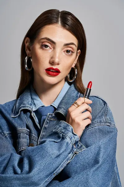 Una joven mujer caucásica en una chaqueta de jean felizmente sostiene un lápiz labial, encarnando estilo y elegancia. - foto de stock