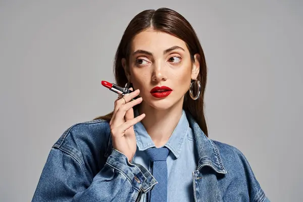A young Caucasian woman with brunette hair in a denim jacket holding red lipstick — Stock Photo