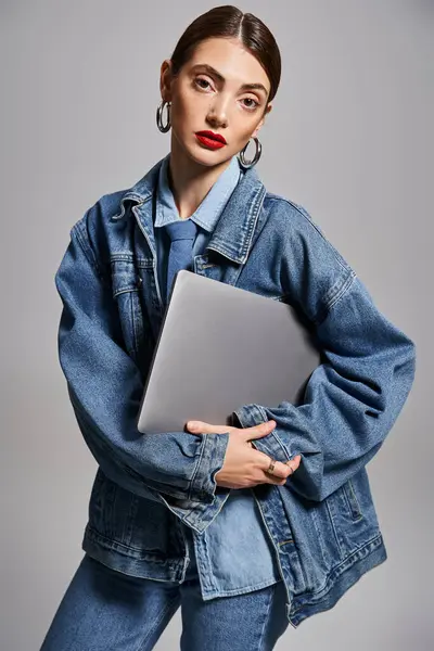 Una joven mujer caucásica con cabello moreno sostiene con confianza un portátil mientras usa una elegante chaqueta de jean en un entorno de estudio. - foto de stock