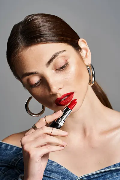 A young Caucasian woman in a denim dress holding a vibrant red lipstick. — Stock Photo