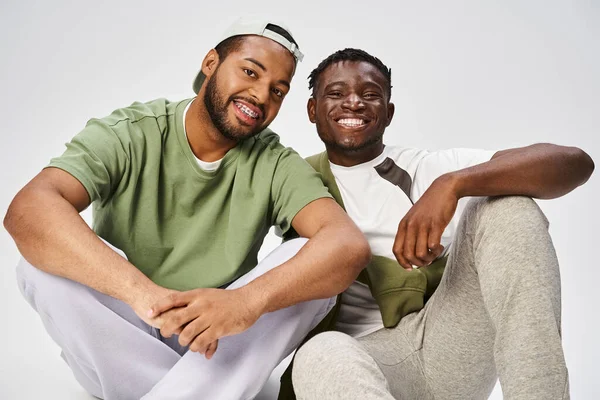 Fröhliche Junifeier, junger afrikanisch-amerikanischer Mann sitzt mit männlichem Freund auf grauem Hintergrund — Stockfoto