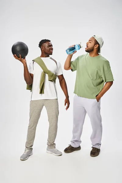 Heureux homme afro-américain tenant ballon et regardant ami boire de l'eau sur fond gris — Photo de stock