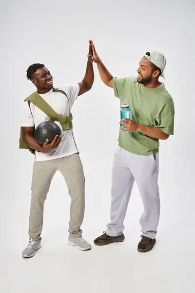 Heureux afro-américains amis masculins tenant ballon et de l'eau tout en donnant haute cinq sur fond gris — Photo de stock
