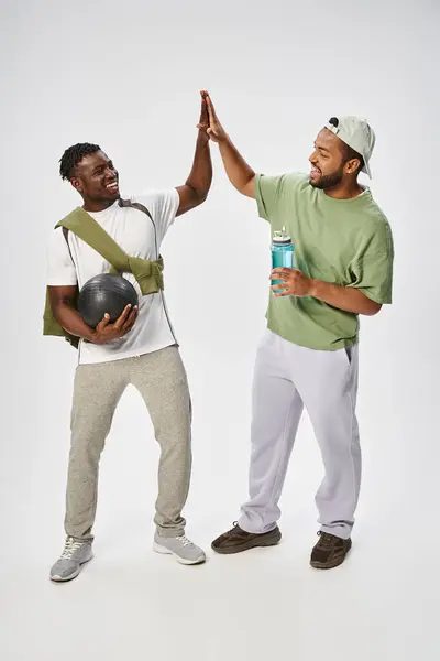 Happy african american male friends holding ball and water while giving high five on grey backdrop — Stock Photo