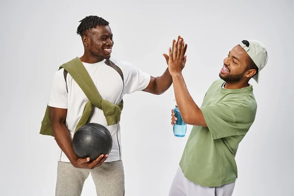 Joyful african american male friends holding ball and water while giving high five on grey backdrop — Stock Photo