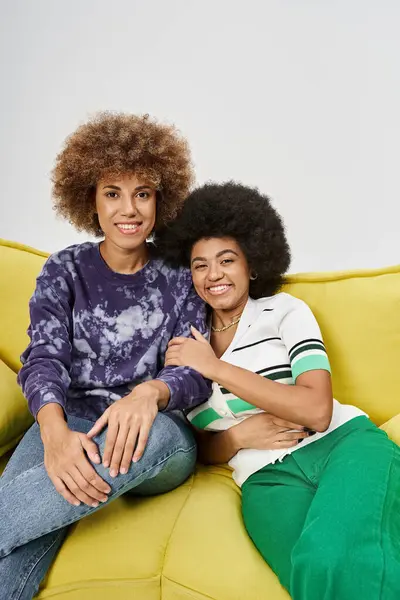 Cheerful and curly african american female friends sitting on yellow sofa on grey background — Stock Photo