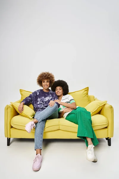 Joyful and curly african american female friends sitting on yellow sofa on grey background — Stock Photo