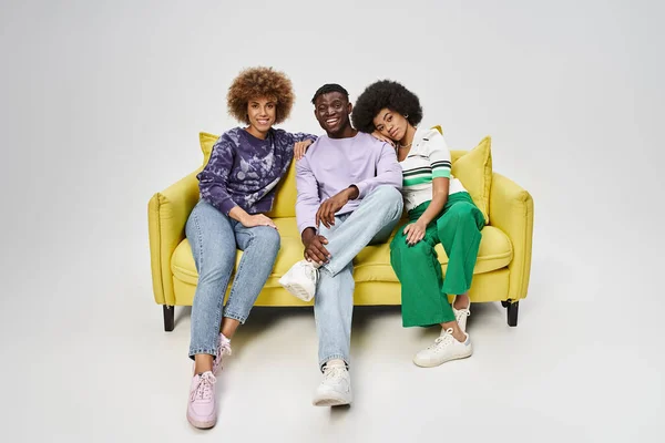 Joyful and curly african american friends together sitting on yellow sofa on grey background — Stock Photo