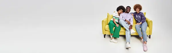 Amici afroamericani gioiosi e ricci insieme seduti sul divano giallo su sfondo grigio, banner — Foto stock