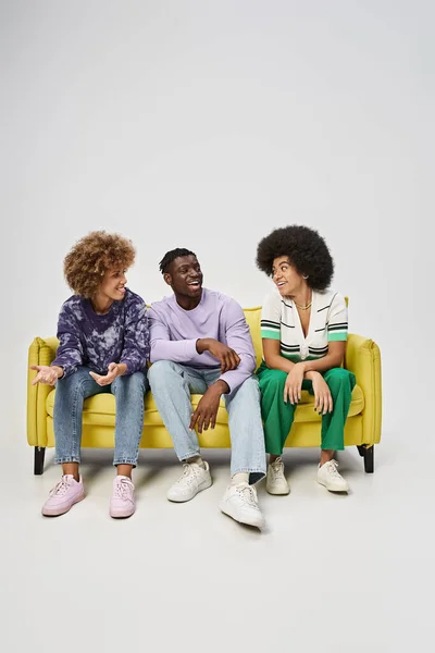 Amigos americanos africanos alegres e encaracolados conversando e sentados no sofá amarelo em fundo cinza — Fotografia de Stock