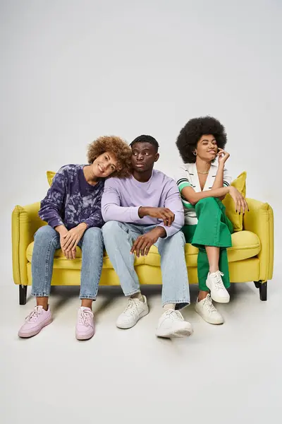 Amis afro-américains joyeux et bouclés se détendre tout en étant assis sur un canapé jaune sur fond gris — Photo de stock