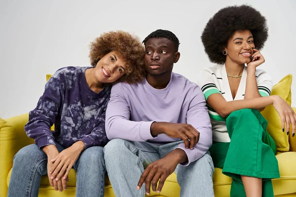 Positive and curly african american friends relaxing while sitting on yellow sofa on grey background — Stock Photo