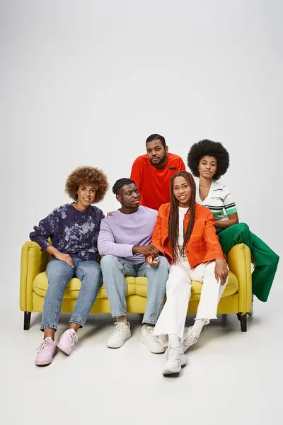 Smiling african american friends in colorful casual attire sitting together on yellow couch on grey — Stock Photo