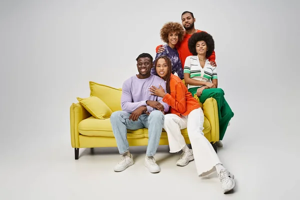 Heureux afro-américains en tenue décontractée lumineuse assis ensemble sur le canapé jaune sur gris — Photo de stock