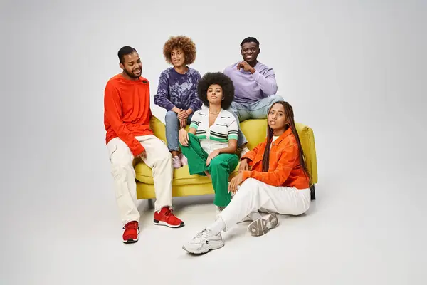 Positive african american friends in bright casual wear sitting together on yellow couch on grey — Stock Photo