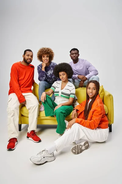 Jolly african american community in bright casual wear sitting together on yellow couch on grey — Stock Photo