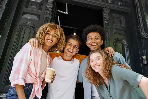 Excited and stylish multicultural buddies hugging and smiling at camera near hostel on city street — Stock Photo