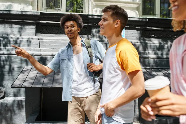 Jovem afro-americano homem apontando com o dedo perto alegre multiétnico amigos andando na cidade — Fotografia de Stock