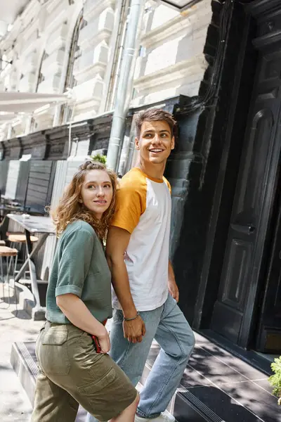 Young and stylish couple of students looking away while entering hostel on urban street, excitement — Stock Photo