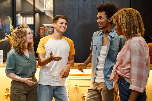 Junger lächelnder Mann im Gespräch mit stilvollen multiethnischen Kumpels in der Lobby der Jugendherberge, Reisende — Stockfoto