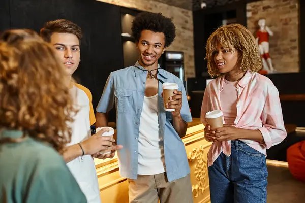 Joyful multiethnic friends with paper cups talking at reception desk in youth hostel, travelers — Stock Photo