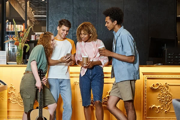 Estudiante mostrando smartphone a amigos multiétnicos con café para llevar y guitarra en el vestíbulo del albergue - foto de stock
