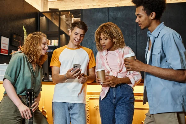 Homme heureux montrant smartphone à des compagnons multiculturels avec café à emporter et guitare dans le hall de l'auberge — Photo de stock