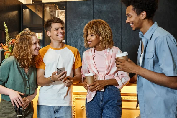 Joven con teléfono inteligente sonriendo cerca de amigos multiétnicos con estilo en la recepción en albergue moderno - foto de stock