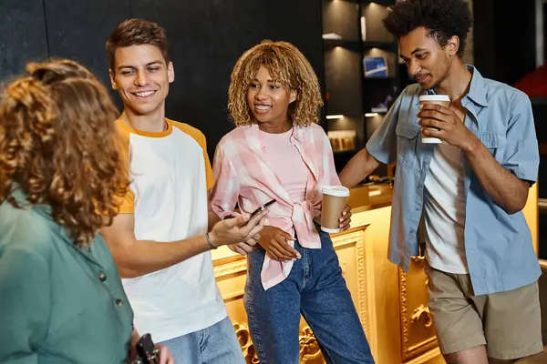 Handsome young man with mobile phone near smiling multiethnic buddies in lobby of modern hostel — Stock Photo
