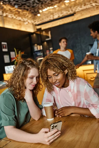 Mujer feliz navegando por las redes sociales en el teléfono inteligente cerca de la amiga afroamericana en el vestíbulo cafetería - foto de stock