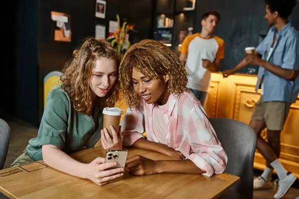 Interracial girlfriends browsing internet on smartphone near coffee to go in cafe of hostel lounge — Stock Photo