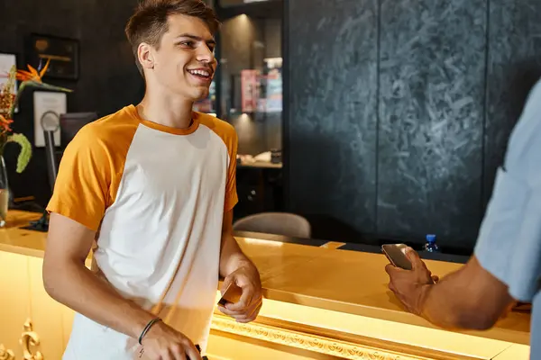 Young smiling man talking to african american friend with smartphone at reception of youth hostel — Stock Photo
