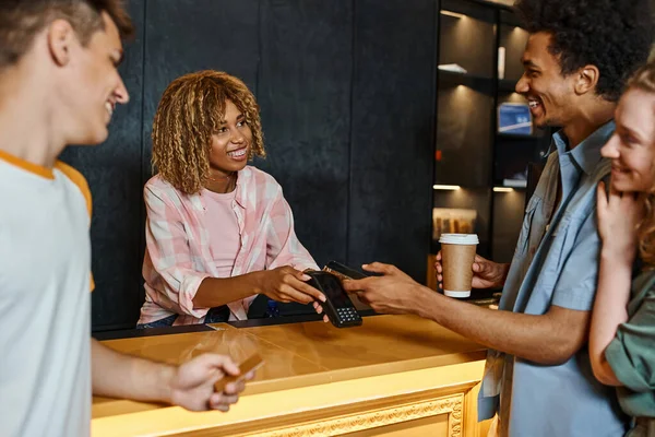 Joyeuse réceptionniste afro-américaine avec terminal de paiement près d'amis multiethniques en auberge — Photo de stock