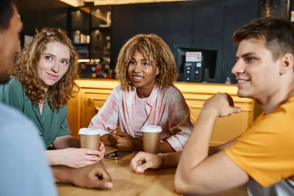 Cheerful multicultural friends talking near coffee to go in lounge cafe of youth hostel, travelers — Stock Photo