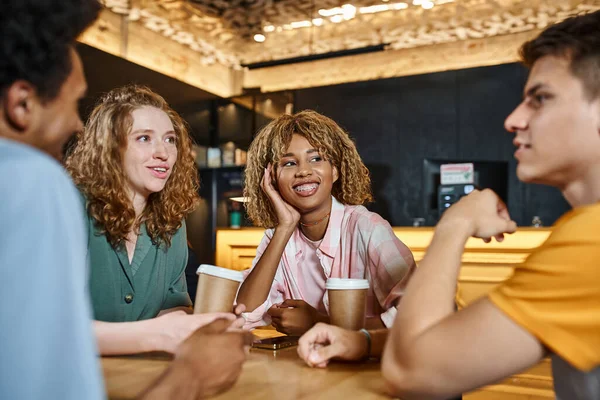 Estudiantes multiétnicos alegres hablando cerca de vasos de papel en salón cafetería de albergue juvenil, amistad - foto de stock