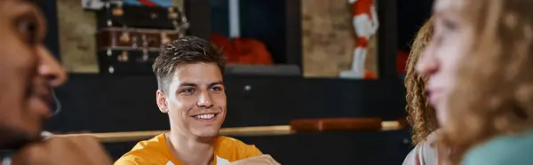 Junger stilvoller Mann lächelt neben multikulturellen Freunden, die in der Lobby des Studentenwohnheims reden, Transparent — Stockfoto