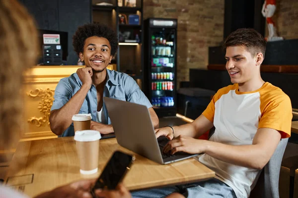 Молодий усміхнений чоловік спілкується на ноутбуці поблизу мультикультурних друзів у фойє кафе студентів гуртожитку — стокове фото