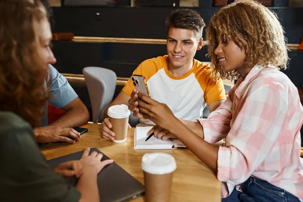 Alegre mujer americana mostrando teléfono inteligente a un amigo sonriente mientras está sentado en la cafetería del albergue, viajeros - foto de stock