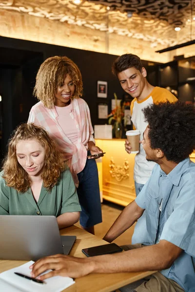 Young woman using laptop near cheerful multicultural friends in lounge cafe of youth hostel — Stock Photo