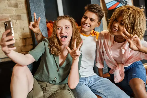 Excited multicultural friends showing victory signs and taking selfie on smartphone in hostel lobby — Stock Photo