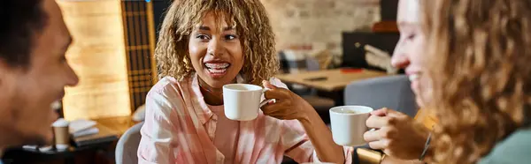 Mulher americana africana alegre com xícara de café perto de amigos multiétnicos no café albergue, bandeira — Fotografia de Stock
