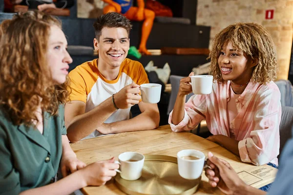 Joyful multicultural friends holding coffee cups and talking in hostel cafe, travel and friendship — Stock Photo